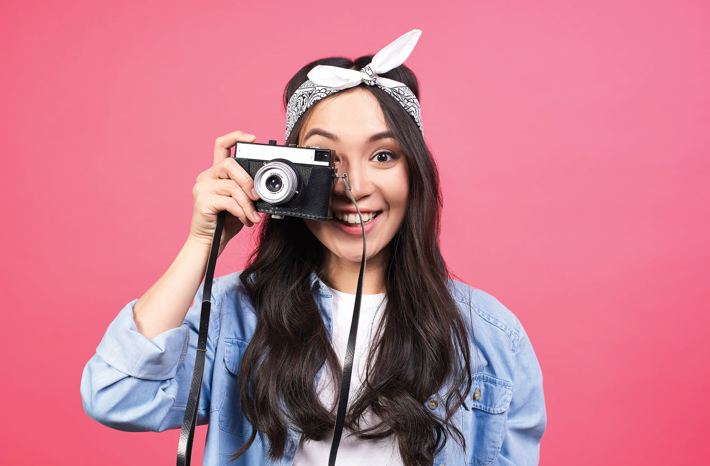Young woman with long hair wearing a bandanna takes a photo with a vintage camera against a pink background.